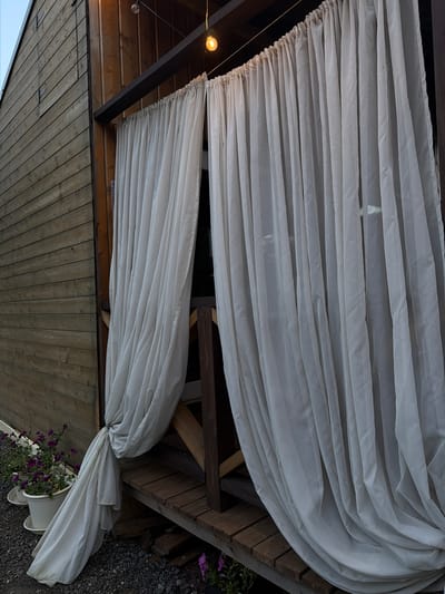 The image shows a wooden structure with white curtains hanging in front of an entrance. A string of lights hangs overhead, with one bulb illuminated. There are two white pots with purple flowers placed on the ground to the left of the structure. The scene appears to be outdoors, possibly in a yard or garden. The location of Kuliki, Russia is not depicted in the image.