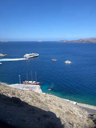This is an aerial shot looking down on a coastal scene in the Thira Municipal Unit, Greece. The image showcases a deep blue, sunlit ocean meeting a rugged, rocky coastline. Several boats are visible in the water and docked.

Dominating the foreground is a large, wooden sailing ship with three masts, docked at a concrete pier. Orange life preservers are visible on its deck, and several people appear to be on board or disembarking. A smaller, modern white vessel with the text "SEARLS" prominently displayed on its side is moving away from the pier, leaving a white wake behind it. Further out in the ocean, a few other smaller boats, some appearing to be pleasure crafts, are scattered across the water. In the distance, the coastline continues, with more rocky cliffs and the vast expanse of the Aegean Sea under a clear, bright sky. The lighting suggests it is daytime, likely mid-morning or afternoon, with the sun casting shadows on the cliff face. The overall scene is one of a serene, sunny day at sea, with maritime activity taking place.