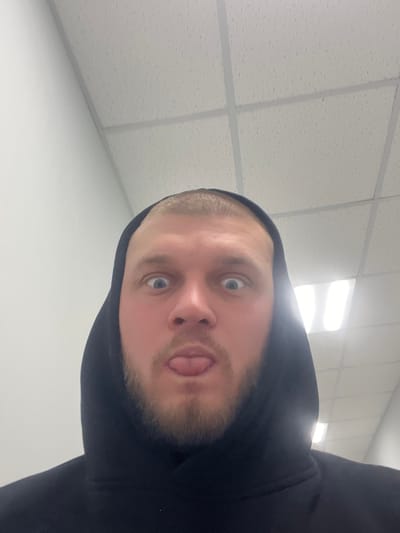 A man with blue eyes and a short blonde haircut wears a black hoodie and sticks his tongue out, peering down at the camera. He has a light beard. The photo appears to have been taken from a low angle, pointing upward at the subject. The background shows a white ceiling with square tiles and fluorescent lighting fixtures.