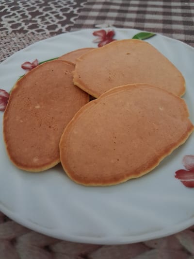A close-up shot shows three golden-brown pancakes arranged on a white plate with a floral pattern. The pancakes are slightly overlapping, with the largest one in the foreground. The plate rests on a patterned tablecloth with brown and beige squares and a floral motif. The background is out of focus. There are no people visible in the image. The scene appears to be indoors, possibly in a home setting in Krolevets, Ukraine.