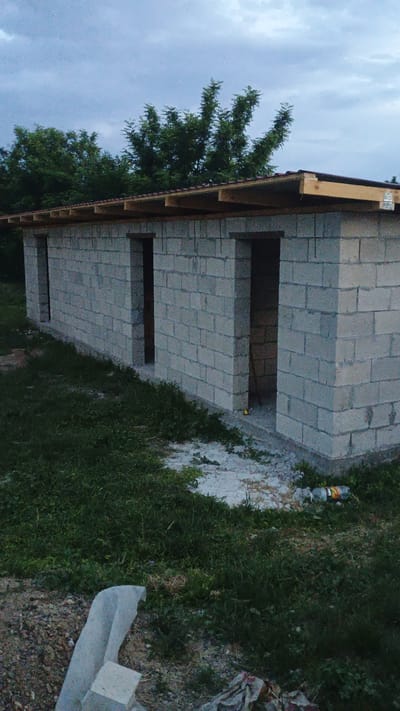 This image shows the side of a cinder block building under construction. The building has a wooden roof structure and several openings for doors or windows. Trees with green foliage are visible on the left side of the building, and the ground is covered in grass, dirt, and small stones. A plastic bottle is lying on the ground near the building. The sky is cloudy, suggesting overcast weather. The scene appears to be a rural or semi-rural location in Mitoc, Moldova, likely during the daytime given the lighting. There is no visible text in the image.