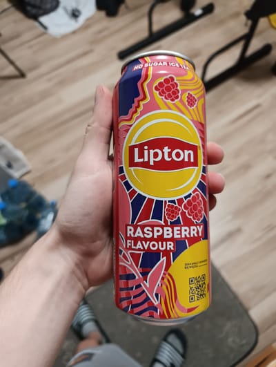 A hand holds a Lipton Raspberry Flavor No Sugar Ice Tea can. The can is predominantly pink with yellow, blue, and red graphics, featuring raspberries and wavy lines. The Lipton logo is prominently displayed in the center. To the right of the can, a QR code is visible, along with Polish text that appears to be an instruction to scan for more information. The background consists of a wooden floor and what looks like furniture legs. In the blurred background, several plastic water bottles are visible. The image appears to be taken indoors, possibly in a home setting. The person's feet, wearing sandals, are also visible at the bottom of the frame.