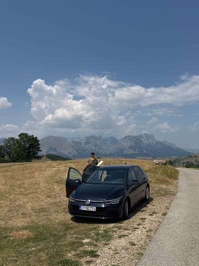 In the city of Pišče, Montenegro, a black Volkswagen Golf is parked on the side of a rural road. The car's driver's side door is open, and a person wearing a green t-shirt and dark pants is standing next to it, holding a piece of white paper and a sandwich. The car is facing the viewer, with its front license plate visible, reading "CT BC774". The surrounding environment is characterized by dry, golden grass, rolling hills, and a dramatic backdrop of rugged, grey mountains under a bright blue sky with scattered white clouds. A few trees are visible to the left of the car. In the distance, on the right side of the frame, a flock of sheep can be seen grazing. The scene suggests a scenic stop during a road trip.