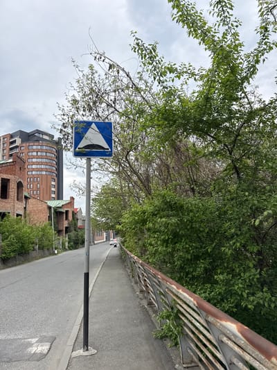 The image shows a road in Assinovskaya, Russia. A blue and white road sign is visible on the left side of the road, partially obscured by tree branches. A metal railing runs along the right side of the road. Buildings are visible in the background, including a multi-story brick building. The sky is overcast. The road surface is asphalt.