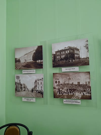 This media file displays an exhibit on a light green wall, featuring four historical black and white or sepia-toned photographs, likely from Borok, Russia. Each photo is under a transparent protective layer and accompanied by a caption.
The top-left photograph shows a multi-story wooden building with many windows, possibly a school, with trees visible around it. Its caption below reads: "Здание женской гимназии 1905 г." (Building of the Women's Gymnasium 1905).
The top-right photograph depicts a large group of people, predominantly young women, arranged in rows posing outdoors in front of a grand, ornate building. The caption to its right states: "Коллектив женской гимназии с Почетным попечителем П.Л. Башениным 1908 г." (Collective of the Women's Gymnasium with Honorary Trustee P.L. Bashenin 1908).
The bottom-left photograph features a long, multi-story building with a light-colored facade and numerous windows, surrounded by trees. The caption below it says: "Здание Алексеевского реального училища Конец ХІХ в" (Building of the Alekseevsky Real School, End of 19th century).
The bottom-right photograph shows a street scene from an early 20th-century town. Buildings line the street, with utility poles and wires visible, and a few individuals can be seen walking. The caption to its right reads: "Здание городского 4-х классного училища (слева) Начало ХХ в." (Building of the city 4-class school (left), Early 20th century), indicating a specific building within the street view.
The overall presentation suggests a historical exhibition, showcasing architectural and social aspects of Borok from the late 19th to early 20th centuries. A black chair with a yellow seat is partially visible on the far right.