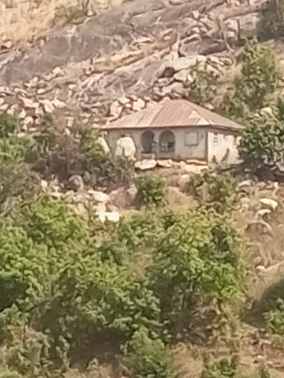 A small, weathered house sits nestled into a rocky hillside. The structure features a simple, rusty roof, arched openings, and faded white walls. Lush green bushes and trees surround the house, contrasting with the rough, light-colored rock formations behind it. The house appears isolated and integrated into the natural environment.