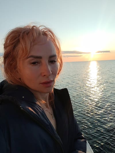 A selfie taken outdoors by a woman with ginger hair and fair skin, wearing a dark jacket with a zipper, and a silver necklace. She is positioned on the left side of the frame, facing slightly to the right. The background features a body of water with sunlight reflecting on its surface, and a bright sky with a few clouds, suggesting a sunset or sunrise. The scene appears to be set at a waterfront, possibly a port or embankment, overlooking a sea or large lake. The location is specified as Kakkarovo, Russia.