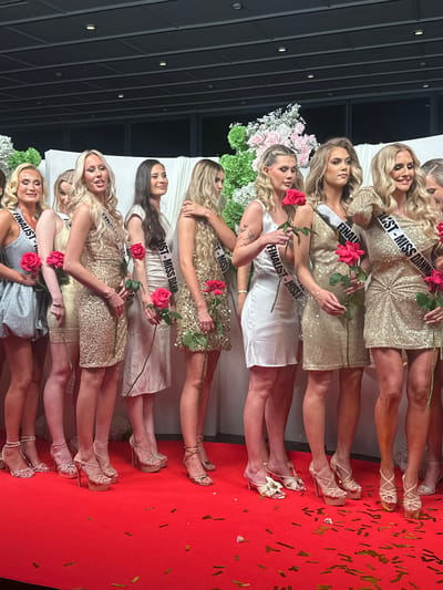 The image depicts a group of women lined up on a red carpet, apparently at a beauty pageant event. The women are dressed in formal dresses, some in gold sequins, one in silver and one in white. They are holding red roses. Each woman is wearing a sash that reads "Finalist - Miss Denmark". Gold confetti is scattered across the red carpet. The venue appears to be well-lit, possibly an event hall.

Given the sash information, it is highly likely that the location is in Skovshoved, Denmark, where the Miss Denmark pageant is often held, though there is no corroborative information in the image. The photo does not allow for any personal identification of the subjects, and there is no further evidence of interaction.