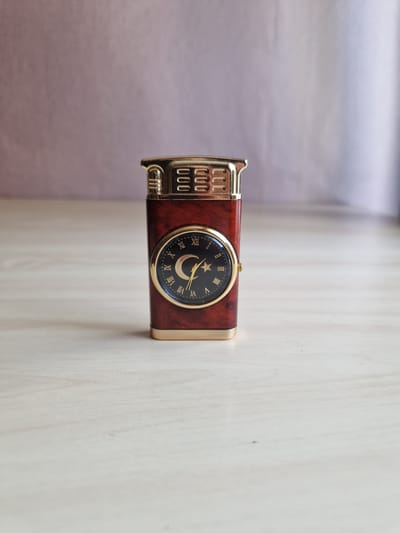 An ornate, rectangular lighter with a dark red wood-like finish and gold accents sits on a light-colored wooden surface. The lighter features a circular clock face with Roman numerals and a crescent moon and star design. The background is a soft, out-of-focus purple-gray fabric. This image is not located in Rikava, Latvia.