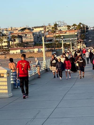 This image captures a daytime scene on the Manhattan Beach Pier in Manhattan Beach, California, USA. The pier is bustling with people walking in both directions. In the foreground, a person wearing a red Cristiano Ronaldo jersey with the number 7 is walking away from the camera. To their left, another person, shirtless and wearing striped pants, stands by the railing. Further down the pier, a diverse group of individuals is seen, some walking together, others in conversation. The background reveals the sandy beach with people enjoying the day, palm trees, and a line of coastal houses and businesses. The sky is clear and blue, indicating good weather. The scene is populated with a variety of people, suggesting a popular recreational area.