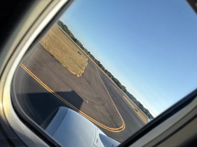 A view from an airplane window shows a tarmac with yellow lines, patches of grass, and a glimpse of trees in the distance. Part of the airplane's engine is visible in the upper right corner of the frame. The sky is light blue and mostly clear. The image is slightly angled, with the horizon at a diagonal. The focus is on the ground below, capturing the textures of the runway and surrounding landscape.