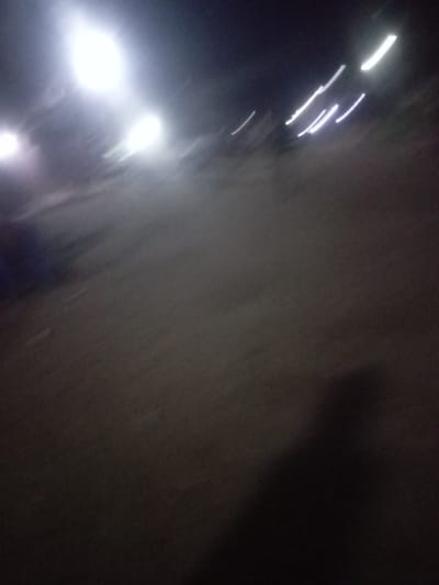 The image depicts a blurry, low-light outdoor scene captured at night in Iyiowa Odekpa, Nigeria. The primary subjects are various light sources and a ground surface.
In the upper left quadrant, three prominent, bright, circular blurs indicate intense stationary light sources, likely streetlights. Towards the upper right, multiple elongated, diagonal streaks of white light suggest moving light sources, possibly from vehicles, or the result of camera motion during a longer exposure.
The central and lower portions of the image show a dark, textured ground surface, appearing to be an unpaved road or path, consistent with the location context. This surface is indistinct due to the low light and motion blur. A large, dark, amorphous shadow occupies the lower right corner, suggesting the presence of a person or object very close to the camera, possibly the photographer's own shadow.
No specific activity or event is clearly discernible, but the pervasive motion blur throughout the image implies either camera movement, the movement of subjects, or both. The time of day is clearly night, characterized by the dark background and the reliance on artificial light. There are no visible details regarding weather, clear human figures, or any discernible text. The overall impression is of a nocturnal environment, possibly a street or pathway, with ambient artificial lighting.
