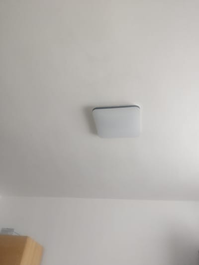 The image depicts a white wall with a single, light-colored square object mounted on it. The object appears to be a device, possibly a sensor or a smart home accessory, with rounded corners and a subtle dark edge. To the right of the object, a corner of a light-colored wooden cabinet or shelf is visible, and below it, a white electrical outlet can be seen. The overall setting is an interior space, likely a room in a building, with a plain white wall dominating the frame. The lighting suggests an indoor environment, possibly during the daytime, though no windows or external light sources are visible. There is no text present in the image. The scene is simple and uncluttered, focusing on the wall-mounted object and its immediate surroundings. The location context provided, Žďár nad Sázavou, Czechia, does not offer specific visual cues within the image itself.