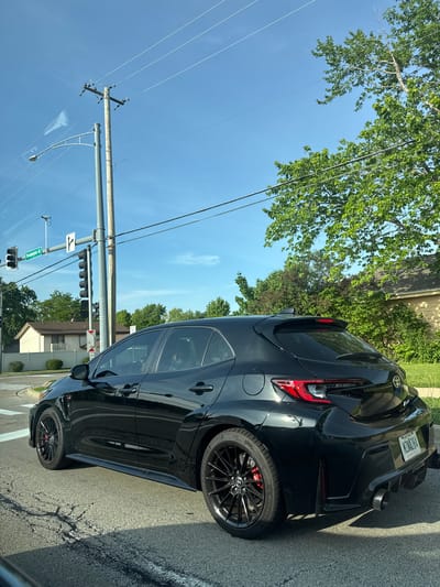 A black hatchback is parked near the corner of the intersection of Bloomingdale Road and W Army Trail Road in Bloomingdale, Illinois. The car has a spoiler and distinctive tail lights. The street sign at the intersection reads "Bloomingdale Rd" and "W Army Trail Rd." A power pole and overhead wires are visible, as are the traffic lights at the intersection. The sky is blue with a few clouds. Trees line the street. The license plate number is N74LB84 and the registered state is Wisconsin.