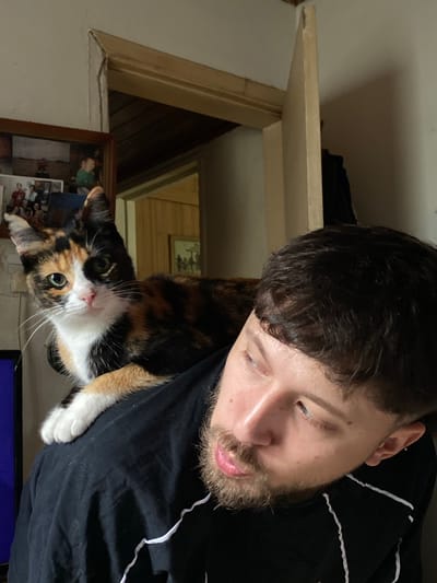 The image presents a close-up, high-angle shot of a man and a calico cat sharing a tender moment in an indoor setting, likely a home in Monte Grande, Argentina. The man, seen from the chest up, has dark hair and a light beard, and is wearing a black shirt accented with white wavy trim. His expression is relaxed, with his gaze directed downwards or to the side, suggesting a moment of quiet contemplation or affection.
Draped over the man's chest and shoulder is a striking calico cat, identifiable by its patches of black, orange/brown, and white fur. The cat's head is turned directly towards the camera, showcasing its bright green eyes and prominent white whiskers, giving it a curious and engaged look. One of its white paws is visible, resting gently on the man's shirt.
The background elements indicate a home environment. A light-colored shelf or wooden surface is visible on the left side, and further back, architectural details like wooden paneling or trim line the walls. A framed picture containing several smaller photographs can also be seen in the lower left background. A hint of a blue screen, possibly a monitor or television, is visible in the bottom right corner. The overall scene conveys a sense of warmth and companionship between the man and his pet. There is no visible text in the image.
