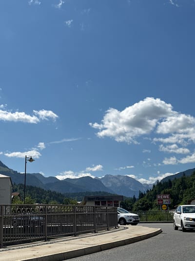 The image shows a street scene in Berchtesgaden, Germany, on a bright, sunny day with scattered clouds. In the foreground, a paved road curves to the right, with a concrete sidewalk and a metal railing to the left. Beyond the railing is a body of water and a street with two cars. One white SUV is parked on the left side of the street, and another white car is driving on the right side of the street.
In the background, there are buildings, including a store with a "Sport Hörhath" sign and a "REWE" supermarket sign with "LIDL" and "dm" logos. Behind the buildings, a lush green forest covers the slopes of majestic mountains, which are prominent in the distance. The sky is a clear blue with fluffy white clouds. A lamppost stands on the left side of the image. The overall impression is a scenic town nestled in the Bavarian Alps.