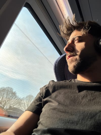 A male individual, estimated to be between 20 and 35 years old, is seated in profile, looking out of a window to his right. He has dark hair, styled upwards, and a beard. He is wearing a plain black short-sleeved t-shirt and black over-ear headphones on his left ear. His mouth is slightly open, and his expression suggests observation. Strong sunlight illuminates the right side of his face and shoulder, casting shadows across his chest and left arm.
The interior environment appears to be a train or bus, indicated by the window structure, the overhead panel, and the blue fabric seat with light polka-dots, which includes a headrest. Through the window, the exterior environment shows a bright, pale blue sky with minimal cloud cover, and the silhouette of several bare, deciduous trees in the mid-distance. Faint horizontal lines, consistent with overhead power lines or reflections, are visible across the upper part of the window. The overall scene is brightly lit by natural light. This scene is located within the city of Ede, Netherlands.