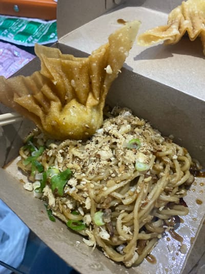 The image shows a close-up of a meal in a cardboard container, likely taken in Walennae, Indonesia. The meal consists of noodles topped with crumbled meat or egg, and chopped green onions. A large, deep-fried wonton is placed on top of the noodles. The container is open, revealing the food. The background is blurred, but appears to show other food packaging and possibly a table. The lighting suggests an indoor setting.