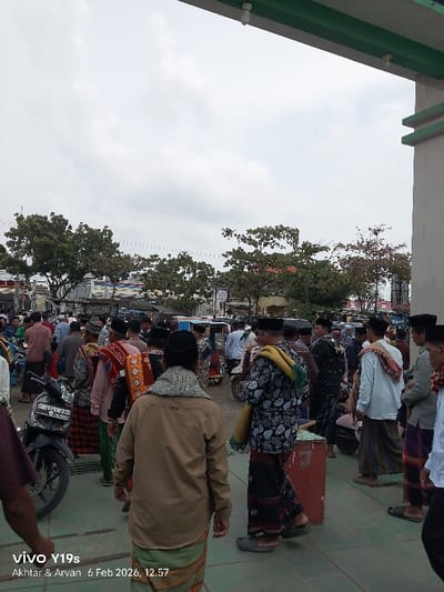A crowded outdoor scene shows people gathered near a building. Many men are dressed in traditional Islamic clothing, including long shirts, sarongs, and caps. A few motorcycles are parked nearby. Trees are visible in the background under a cloudy sky. The architecture suggests a mosque or religious site.