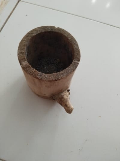 A wooden mortar with a small protrusion at the bottom rests on a white tiled surface. The mortar is cylindrical and hollow, containing a dark substance. The background is a light-colored, tiled floor. The image is taken from a high-angle perspective, looking down at the mortar. The location is identified as Meunasahtunong, Indonesia.