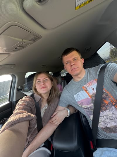 The image shows two individuals, a man and a woman, seated in a car. The man is in a gray t-shirt with a graphic design and is wearing a seatbelt. The woman is wearing a light brown top and is also wearing a seatbelt. The interior of the car is visible, including the dashboard, sun visor, and seats. The location cannot be determined as Lipaki, Russia, without additional context within the image.