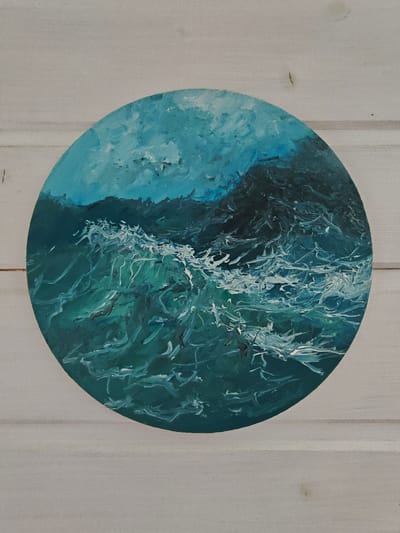 The image is a close-up, top-down shot of a circular painting of rough, turbulent ocean waves, rendered in shades of teal, deep blue, and white. The painting has a textured, impasto style, with thick brushstrokes creating a sense of movement and volume in the water. The waves appear to be breaking, with white foam cresting and swirling. The background of the painting shows darker, more abstract shapes that could represent a distant, shadowy coastline or the ocean depths. The painting is positioned on a light-colored, horizontal wooden surface, likely a wall or table, which shows visible grain and plank lines. There is no indication of the city of Koledino, Russia, or any other specific location within the image itself.