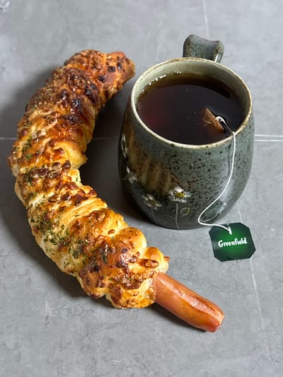 A close-up, eye-level shot shows a baked pastry wrapped around a sausage, placed next to a mug of tea with a tea bag. The pastry is golden brown and appears to be sprinkled with herbs and cheese. The sausage is partially visible, protruding from one end of the pastry. The mug is a speckled green ceramic with a handle and features a delicate floral pattern. A white string from the tea bag dangles into the mug, and a green tag labeled "Greenfield" is attached to the string. The background is a textured, gray surface. The scene appears to be indoors, possibly on a countertop or table. There is no indication of the city of Gwangju, South Korea.