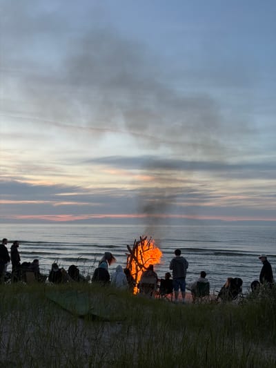 In Plieņciems, Latvia, a group of people are gathered on a sandy beach near the sea at dusk. A large bonfire burns brightly, casting an orange glow on the faces of those around it. Smoke rises from the fire and drifts into the sky, mingling with the soft colors of the setting sun. Several individuals are seated in chairs, facing the fire or the ocean, while others stand and observe the scene. The sea is calm with gentle waves rolling towards the shore. Tall grasses grow along the edge of the beach, adding a natural texture to the foreground. The sky is a gradient of blues and grays, with hints of pink and orange from the sunset.