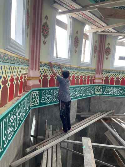 An adult male stands barefoot on a makeshift wooden scaffolding inside a building in Mns Asan, Indonesia, actively engaged in decorative painting. The man, wearing a paint-splattered grey short-sleeved shirt and dark trousers, is seen from the back, reaching upwards with both hands towards a wall. The interior features elaborate Islamic architectural and decorative elements. The upper wall section is light yellow/cream, adorned with ornate pink and red columns. Below a row of arched, open windows, a horizontal band displays repeating geometric patterns in gold, blue, green, red, and brown. Below this, a darker red/brown section exhibits a series of inverted arch-like shapes. A prominent teal-green band with white Arabic calligraphy runs horizontally below the red/brown section, directly above the man's eye level. The lower visible wall section is unfinished concrete. A small, diamond-shaped window is visible on this lower wall. The wooden scaffolding consists of multiple planks and beams, some of which show paint splatters. The bright light streaming through the unpaned arched windows illuminates the scene, indicating an interior under active finishing.