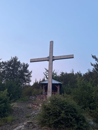 The image depicts a large, metallic cross situated outdoors in a rocky, wooded area. The cross is centrally positioned and appears to be made of concrete or a similar material, with a rough, textured surface. Beneath the cross and slightly to the right, there is a small, open-sided shelter with a pitched roof made of corrugated metal. The shelter has wooden support beams and a floor. The ground leading up to the shelter is covered in loose stones and rocks, with patches of green grass and bushes growing throughout. A stone wall, constructed with irregular stones, is visible behind the path and to the right of the shelter, forming a terraced effect. The background is filled with dense green trees and foliage, characteristic of a forest. The sky above is a clear, pale blue, transitioning to a faint pinkish hue near the horizon, suggesting either dawn or dusk. There are no people visible in the image. The location is identified as Strazha, Bulgaria.