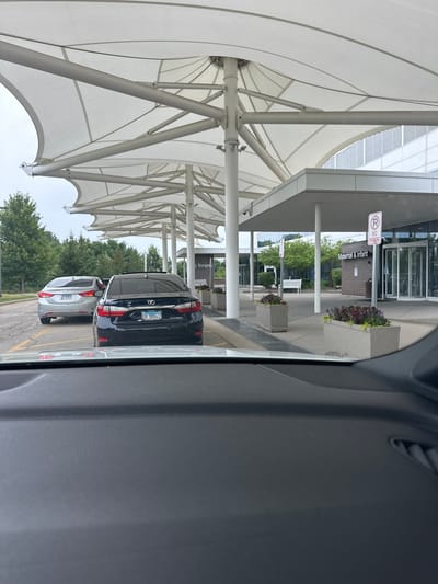 The image depicts the exterior of a hospital building with a large, white, canopy-like structure extending over a drop-off or waiting area. In the foreground, the dashboard of a vehicle is visible. Two cars are parked or waiting in front of the building. The car on the left is silver, and the car on the right is black. The building has a modern architectural design with large glass windows and doors. A sign on the building reads "Maternal & Infant." There are planter boxes with flowers along the walkway. The overall environment appears to be a typical hospital entrance. The location is identified as Hoffman Estates, United States of America.