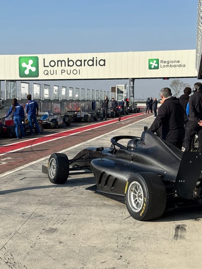 The image displays an outdoor scene at a motor racing pit lane or grid area, located at the Cremona Circuit in San Martino del Lago, Italy. An overhead gantry structure prominently features signs reading "Lombardia QUI PUOI" and "Regione Lombardia," each accompanied by a green stylized "L" logo. The ground surface is light-colored concrete, marked with a distinct red painted line. The sky above is clear and blue.
In the foreground, positioned to the right, the rear and side of a dark carbon-fiber open-wheel race car are visible, oriented away from the camera. This vehicle is equipped with silver multi-spoke wheels and Pirelli P Zero tires bearing yellow text. A man dressed in a dark long-sleeved shirt stands behind this specific race car.
In the mid-ground, a linear arrangement of at least five additional open-wheel race cars are staged on the red-marked lane, also oriented away from the camera. These cars display varied liveries, including elements of red, white, and blue. Approximately ten individuals are present in this section of the scene. On the left, two individuals wearing blue team uniforms are positioned near a red and white race car. Further along the lane, several men in dark clothing, some equipped with headsets, are standing near the parked vehicles and adjacent to the gantry structure. All race cars are stationary, indicating a pre-session or pre-race staging activity.