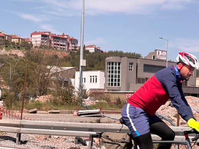 A person wearing a red, white, and blue cycling outfit and a helmet is sitting on a metal railing in Menteşe, Turkey. The person is wearing bright yellow gloves and is resting their arm on the rail. Behind the person, there is a paved road with gravel on the side and green vegetation. There are several buildings in the background, including a gray building with a sign that reads "KIRALIK." Beyond the buildings, there are trees and houses on a hillside. The sky is light blue with a few clouds.