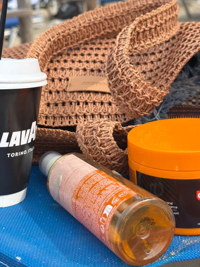 The image is an overhead shot of several personal care items and a beverage on a blue, textured surface, possibly a beach towel. In the foreground and slightly left, a black disposable coffee cup with the "LAVA" logo and "TORINO, ITAL" is visible. To the right of the coffee cup, a clear plastic bottle containing amber liquid lies on its side, with labels displaying text in multiple languages and a "PET" recycling symbol. To the right and slightly behind the bottle is a bright orange jar with a dark label, the top of which is prominently featured. In the background, a woven brown bag, possibly made of straw or raffia, is partially visible, its textured weave creating a focal point. The overall setting appears to be outdoors, with natural light illuminating the scene. The location is the city of Olympiada, Greece.