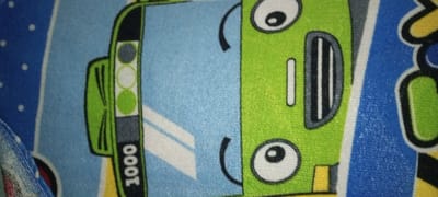 A patterned fabric depicts a cartoon bus. The bus is green with large, round eyes and a smiling grill. It has traffic light icons on the side, with the number "1000" below. The bus is set against a light blue background. Part of another design is seen on the right, featuring blue, white, and yellow colors. The image has an overall soft, textured look.