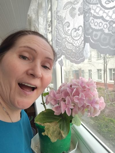 A woman is captured in a close-up selfie indoors, standing beside a window with a vibrant pink potted plant. She has brown hair, fair skin, and is wearing a blue top with a silver necklace. Her mouth is wide open in what appears to be an expression of surprise or excitement, and she is looking directly at the camera.
The plant, which resembles a hydrangea, features clusters of delicate pink blooms and is situated in a green pot on the windowsill. Some of its leaves show signs of yellowing and browning. White lace curtains with intricate floral and scroll patterns hang over the top half of the window, diffusing the natural daylight.
Beyond the window, a multi-story white brick building with numerous rectangular windows is visible. Several bare trees stand in the foreground of the building, suggesting a cool season like late autumn, winter, or early spring. The ground beneath the trees appears somewhat barren, and a metal fence is also discernible. The scene takes place in Yanaul, Russia. There is no visible text in the image.