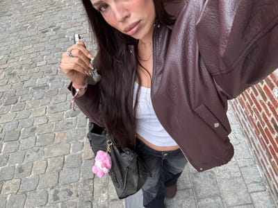 A high-angle image captures a female individual standing on a cobblestone street in Tielrode, Belgium. The individual is partially visible, oriented facing the camera, with long dark hair. She wears a dark brown faux leather bomber jacket, unzipped, over a white ribbed crop top, paired with dark grey or black patchwork-style trousers. Dark footwear is partially visible on the ground. The individual's right hand holds a set of keys with a silver-toned keychain. A silver-toned ring is present on her right ring finger, and a red and white braided bracelet is on her right wrist. Fingernails exhibit a light-colored polish. A dark-colored shoulder bag, from which a pink, fluffy, animal-shaped keychain hangs by a gold-colored chain, is carried by the individual. The ground surface consists of grey, irregularly shaped cobblestones. A red brick wall with visible mortar lines forms the background element on the right side of the frame.