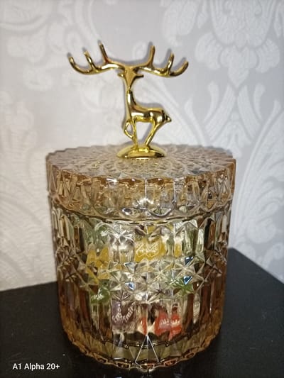 A gold-colored deer figurine is perched atop a decorative, faceted glass jar filled with colorful, foil-wrapped candies. The jar has a textured, diamond-patterned surface and is amber-toned. The background features a light-colored wallpaper with a subtle floral or damask pattern. The jar is resting on a dark, flat surface, casting a shadow to its right. The image is an indoor still life, likely taken in a domestic setting within Улукаўскі сельскі Савет, Belarus.