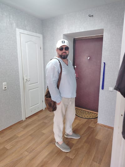 A man stands in what appears to be an apartment hallway in Malaryta, Belarus. He is wearing a light blue, long-sleeved collared shirt, loose-fitting beige pants, gray slip-on shoes, a white baseball cap, and dark sunglasses. A brown leather messenger bag is slung over his left shoulder. The man has a beard and is looking towards the camera.
The hallway has gray textured wallpaper on the walls. To the left of the man is a white door with a silver handle. To his right is a dark brown, metal-clad door with a silver peephole and handle, suggesting it's an entrance door. A circular doormat with a geometric pattern is placed in front of the dark door. On the wall to the right of the dark door, a blue shoehorn hangs vertically next to a light switch. The floor is made of light brown wooden laminate. A light gray electrical cord runs across the floor in front of the man. The lighting suggests it is daytime, and there are no indications of weather. The overall mood appears neutral, with the man posing for a photo. There is a small white logo on the front of the man's baseball cap, but the text is not legible.
