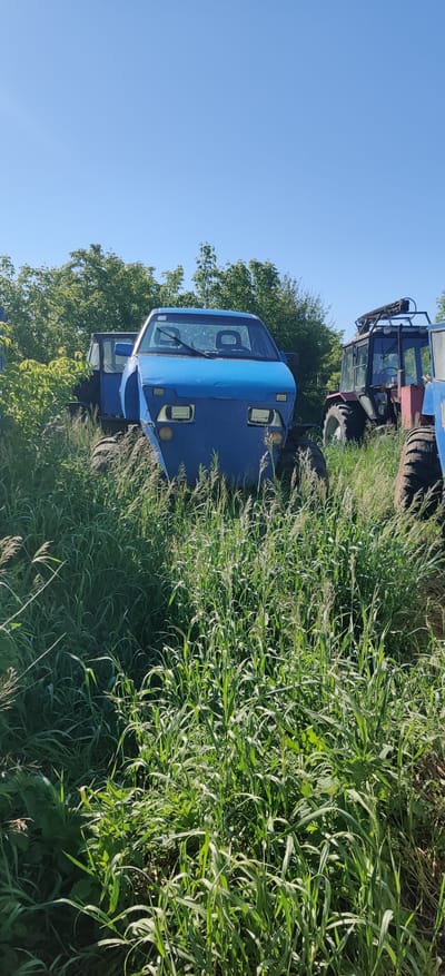 The image, taken in Bezpalivka, Ukraine, depicts an outdoor scene on a bright, clear day, likely in a rural or agricultural setting. The sky is a cloudless, vibrant blue.
The primary subjects are two large agricultural vehicles, possibly tractors or specialized farm equipment. In the foreground, positioned slightly to the left of the center, is a sturdy blue vehicle facing the viewer. It features a prominent cabin with a large windshield and wipers. Rectangular LED-style headlights are visible on its front panel, which has a textured or perforated surface. Below the windshield, there's a visible white detail, possibly a part of its structure or a marking.
Behind and to the right of the blue vehicle, a second piece of heavy machinery is partially visible. This vehicle appears to be red and blue, with large, rugged tires and a cabin section similar to the first vehicle. Both vehicles seem to be parked or stationary amidst abundant vegetation.
The ground is covered with tall, lush green grass and possibly wild plants, indicating an overgrown area or a field. In the background, a line of green trees provides a natural backdrop. The bright sunlight casts distinct shadows, highlighting the clear weather conditions. There is no discernible activity taking place, and no visible text can be clearly read on any part of the vehicles or environment.