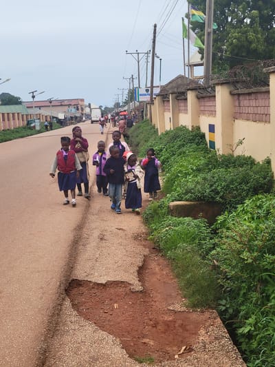 A group of seven children, appearing to be of primary school age, walk along the side of a paved road in Tangu, Nigeria. The children are dressed in school uniforms, consisting of blue skirts or dresses and various tops, including jackets and shirts. One child is carrying a bottle of water. The road surface is uneven with a significant pothole on the right side. To the right of the road, there is a concrete wall topped with barbed wire, and lush green vegetation grows alongside it. In the background, several buildings and utility poles are visible. The sky is overcast.