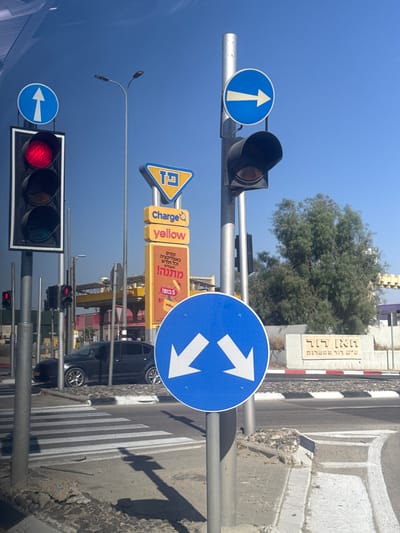 The image, taken in Arad, Israel, shows a street scene with traffic signals and road signs under a clear blue sky, suggesting a sunny day. The perspective is tilted, giving a somewhat disorienting view of the environment.
Prominently featured are several traffic lights. One shows a red light, indicating a stop. Another traffic signal is visible in the distance, with its lights obscured or off. Alongside the traffic signals are various road signs. A circular blue sign with a white arrow pointing upwards indicates a mandatory forward direction. Another circular blue sign displays two white arrows pointing diagonally upwards and to the right, suggesting lanes diverge in those directions. A triangular yellow sign with the letters "פז" (Paz) and a "G" symbol is also visible, likely indicating a gas station or related service. Additionally, there are yellow rectangular signs with Hebrew text, one saying "Charge yellow" and another featuring text that seems to be an advertisement for a gift with purchases made through an app.
A street with a pedestrian crossing is visible, and a dark-colored car is parked or driving near it. In the background, a building with a distinctive facade, possibly with a patterned top, and a large green tree are present. A street sign with Hebrew text, "ראן דוד שיש לול טטריק," is mounted on a pillar. The overall impression is a typical urban street setting during daylight hours.
The image captures a moment of everyday urban life, focusing on the infrastructure that guides traffic and informs the public. The tilted angle and the variety of signs contribute to a dynamic, albeit slightly unusual, visual narrative of the street.
---
**Note on Hebrew Text:**
- The sign "פז" is a prominent Israeli gas station chain.
- The yellow signs appear to be advertisements, with one mentioning "Charge yellow" and another referencing a gift or promotion for app users.
- The sign "ראן דוד שיש לול טטריק" is likely a street name or a local landmark sign. The exact meaning of "שיש לול טטריק" is unclear without further context, but "ראן דוד" could be a street name.