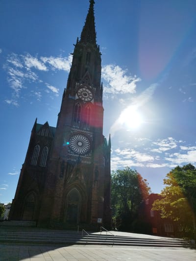 The image shows a low-angle view of the St. Mary's Church (St. Marienkirche) in Bremerhaven, Germany. The towering red brick church with Gothic architectural elements dominates the frame, its spire reaching towards a bright blue sky dotted with white cumulus clouds. The sun is intensely bright, creating lens flare and a visible sunbeam effect in the upper right portion of the image. The church features a prominent clock face and a large rose window. In the foreground, wide stone steps lead up to the church entrance, and paved ground is visible. To the right of the church, green trees and a dark-colored building are partially obscured by foliage. The overall scene suggests a clear, sunny day.
