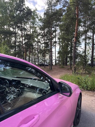 A bright pink Mercedes-Benz C-Class is parked on a dirt road in a wooded area. The car is viewed from the passenger side, with the interior visible through the open door and window. The dashboard features a digital display and climate controls. In the background, several other vehicles are parked among tall pine trees. A body of water is visible in the distance. The scene appears to be a scenic overlook or a picnic spot. The location is identified as Bolshaya Izhora, Russia.