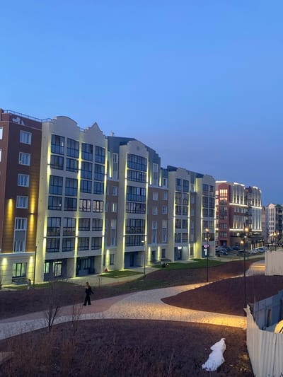 The image depicts an evening scene within a modern residential area, likely in Chemoshur, Russia. The sky is a clear, deep twilight blue, indicating a time after sunset.

Multiple multi-story apartment buildings line the left side of the frame. These structures feature contemporary architecture with facades in various shades of brown, beige, and gray, accented by large, dark-framed windows and numerous balconies. The buildings are brightly illuminated by warm-toned exterior lights mounted along their facades and under balconies, creating a welcoming glow. Some interior lights are also visible through the windows. Faint reflected Cyrillic text, possibly "ЛЕЙБЫ" (labels/brands), is discernible on a ground-floor window of one building.

On the right side, a winding, light-colored paved pathway, possibly concrete or stone, extends into the distance. This pathway is also illuminated by ground-level lights, casting bright pools of light. The ground bordering the path is covered with dark soil or mulch and sparse, bare vegetation, suggesting late autumn or early spring. A single person, dressed in dark clothing, is seen walking away from the viewer on this pathway.

In the foreground on the far right, a temporary corrugated metal fence is visible, alongside a small pile of white snow. In the background, further residential buildings are present, with some parked cars visible between them. The overall impression is one of a peaceful, well-lit urban or suburban residential complex after dusk.