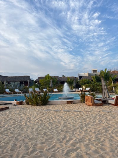 The image depicts a coastal scene, likely a resort, in El Jícaro, Mexico. The view is a sideways-oriented shot capturing a blue and white sky, a sandy beach, and resort buildings alongside a narrow swimming pool. The sky features elongated, streaky clouds. The beach is light beige, marked with patterns typical of dry sand and occasional embedded stone pavers. Adjacent to the beach is a rectangular swimming pool with a visible fountain feature. Thatched-roof structures, likely cabanas, and tropical vegetation such as palm trees, provide cover. Beach chairs and umbrellas are arranged near the pool, implying a relaxed leisure environment. The image does not depict any people or specific actions.