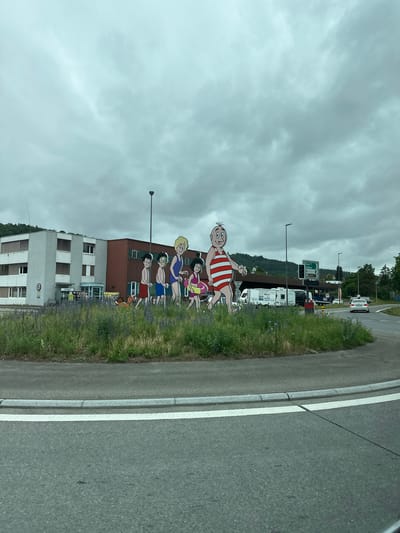 The image shows a roundabout in Zurzach, Switzerland, with a large, colorful cutout of a cartoon family wearing swimwear. The family consists of a man in a red and white striped swimsuit, a woman in a blue swimsuit, and three children. A dog is also depicted. Behind the cutout, there is a modern building with a brown facade and large windows, and to its left, a lighter-colored building with multiple floors and horizontal blinds. A sign on the building reads "VITOGAZ". The roundabout is surrounded by green grass and wildflowers. Traffic signs are visible in the background, indicating directions to Basel and Koblenz. The sky is overcast with grey clouds. A white car is seen in the distance on a road to the right of the roundabout.