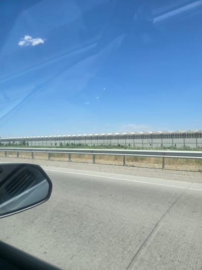 The image depicts a roadside view of a long row of greenhouses, indicative of agricultural activity. These structures, with their distinctive arched roofs, stretch across the horizon under a clear blue sky dotted with a few wispy clouds. A metal guardrail separates the road from the area where the greenhouses are situated, and beyond that, sparse vegetation and dry earth are visible. The foreground shows the asphalt of a road, with the reflection of a car's interior visible in the side-view mirror on the left. The scene appears to be taken from inside a vehicle. There are no people or animals visible in the image. The location is specified as Rabatiohun, Uzbekistan.