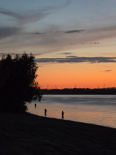 The image depicts a sunset scene at the Zhiguli Sea, a reservoir on the Volga River near Zhigulyovsk, Russia. In the foreground, a dark silhouette of a tree lines the left edge of the frame, casting a shadow over the sandy shore. Three figures, silhouetted against the water, are engaged in fishing. One person stands further back in the water, while the other two are closer to the shore, with one holding a fishing rod. The water's surface reflects the warm hues of the sunset, which transitions from a vibrant orange near the horizon to a soft blue-gray at the top of the frame. The distant horizon is marked by a dark line of trees, punctuated by industrial towers and lights, indicating the presence of civilization. The sky is filled with scattered clouds, some tinged with the colors of the setting sun.