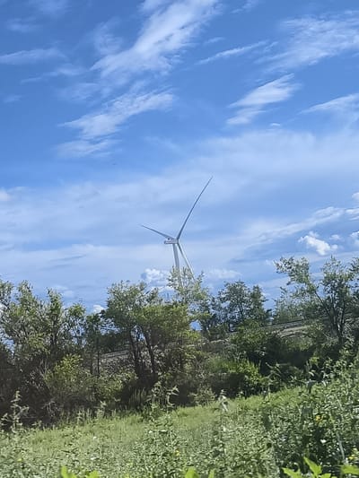The image captures a scenic view of a wind turbine against a bright blue sky with scattered white clouds. The turbine, a modern structure, has two prominent blades extended at an angle, suggesting it is either stationary or slowly rotating. Below the turbine, a dense line of green trees and foliage forms a natural barrier, with a hint of what appears to be a railway track running through them. The foreground is dominated by lush green grass and smaller plants, creating a natural, slightly wild landscape. The overall scene suggests an outdoor setting on a clear, possibly warm day, given the vibrant blue sky and bright lighting. There is no visible text in the image. The location is identified as Ақжар, Kazakhstan, which is consistent with the presence of wind turbines in some regions of the country.
